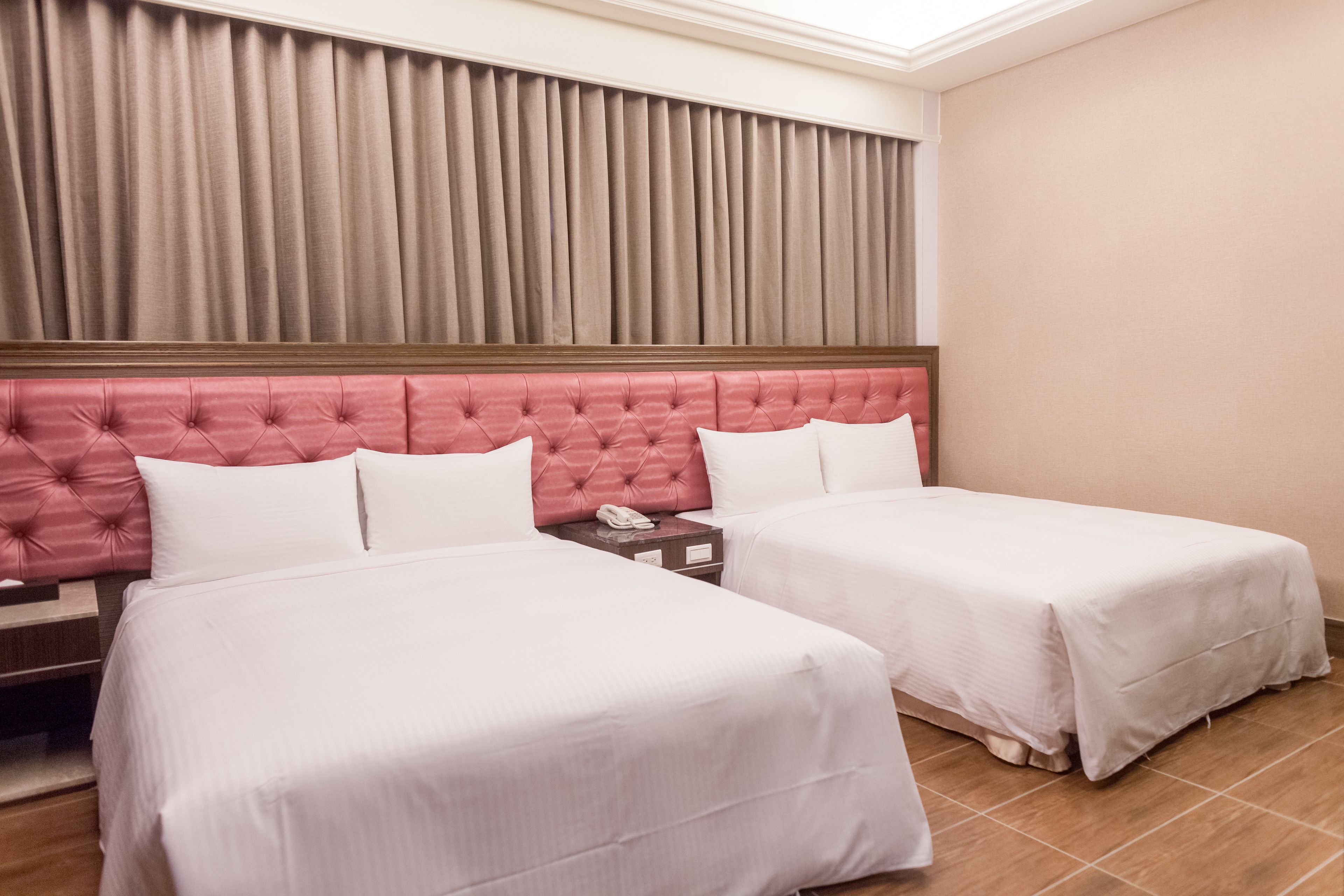 Habitación de hotel con dos camas, cabecera de madera, mesita de noche con teléfono y ventana con cortinas.