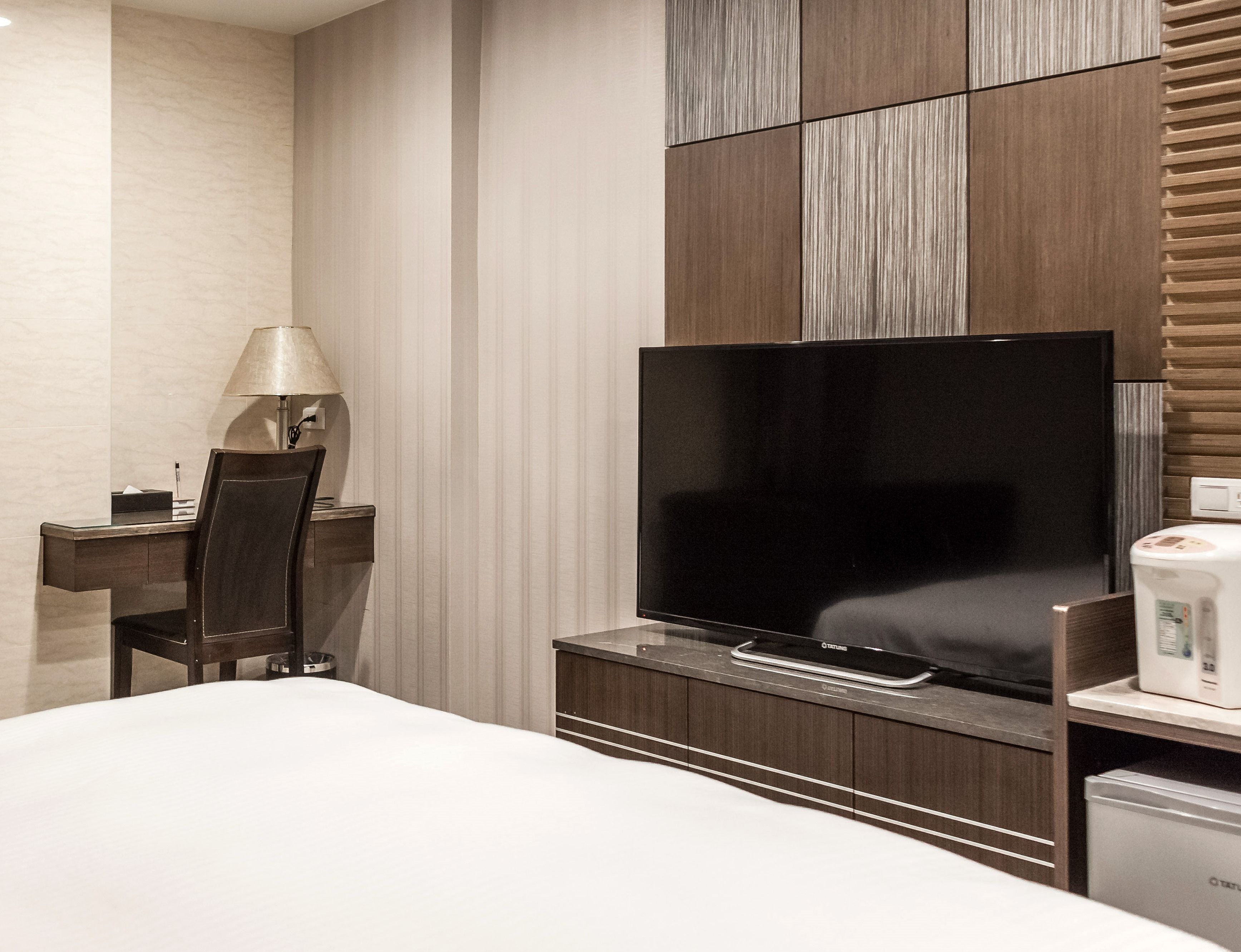 A hotel room with a bed, a desk with a lamp, a flat-screen TV, and a minibar.