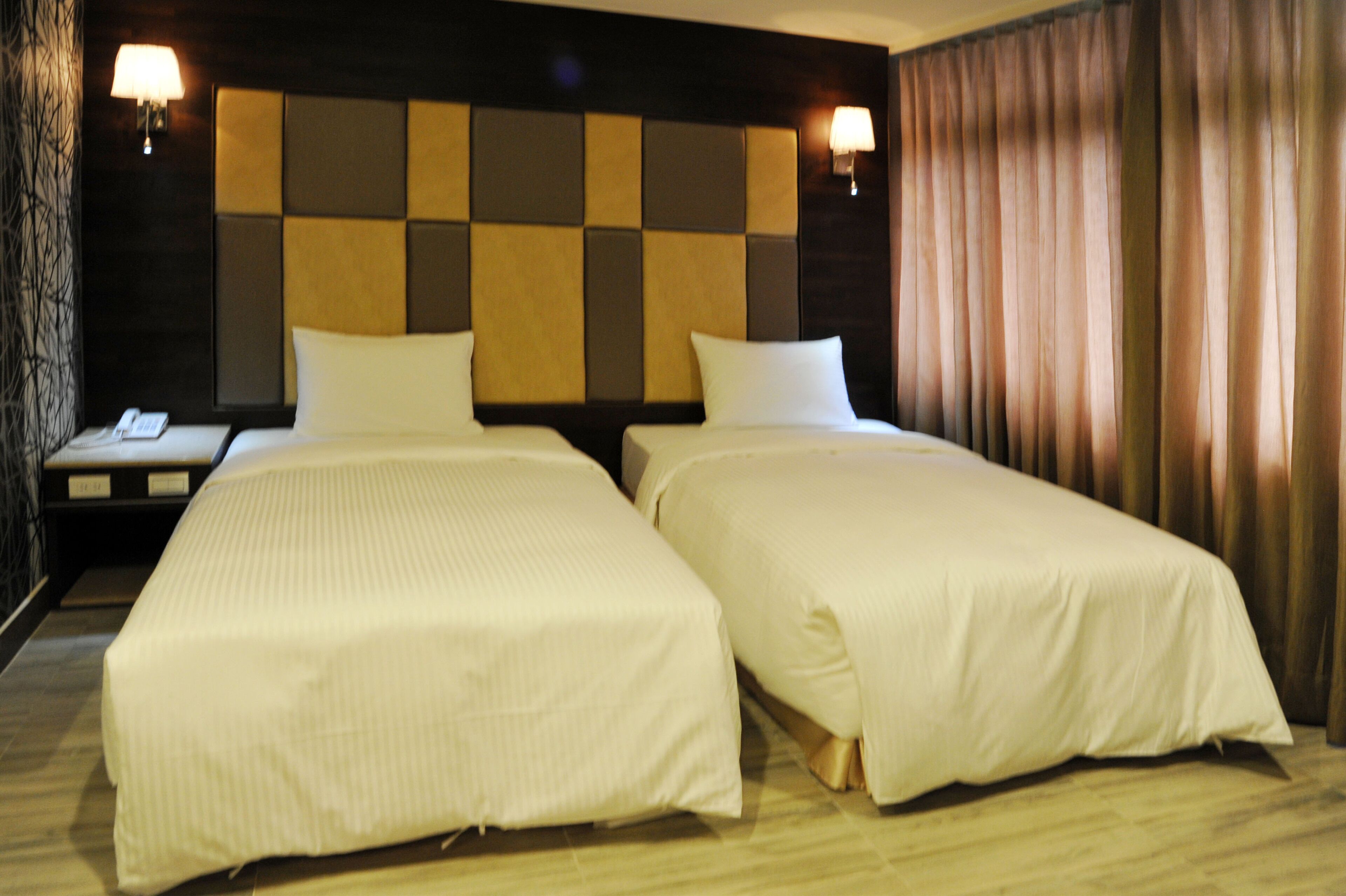 A hotel room with two single beds, a nightstand with a lamp, and a headboard with a square pattern.