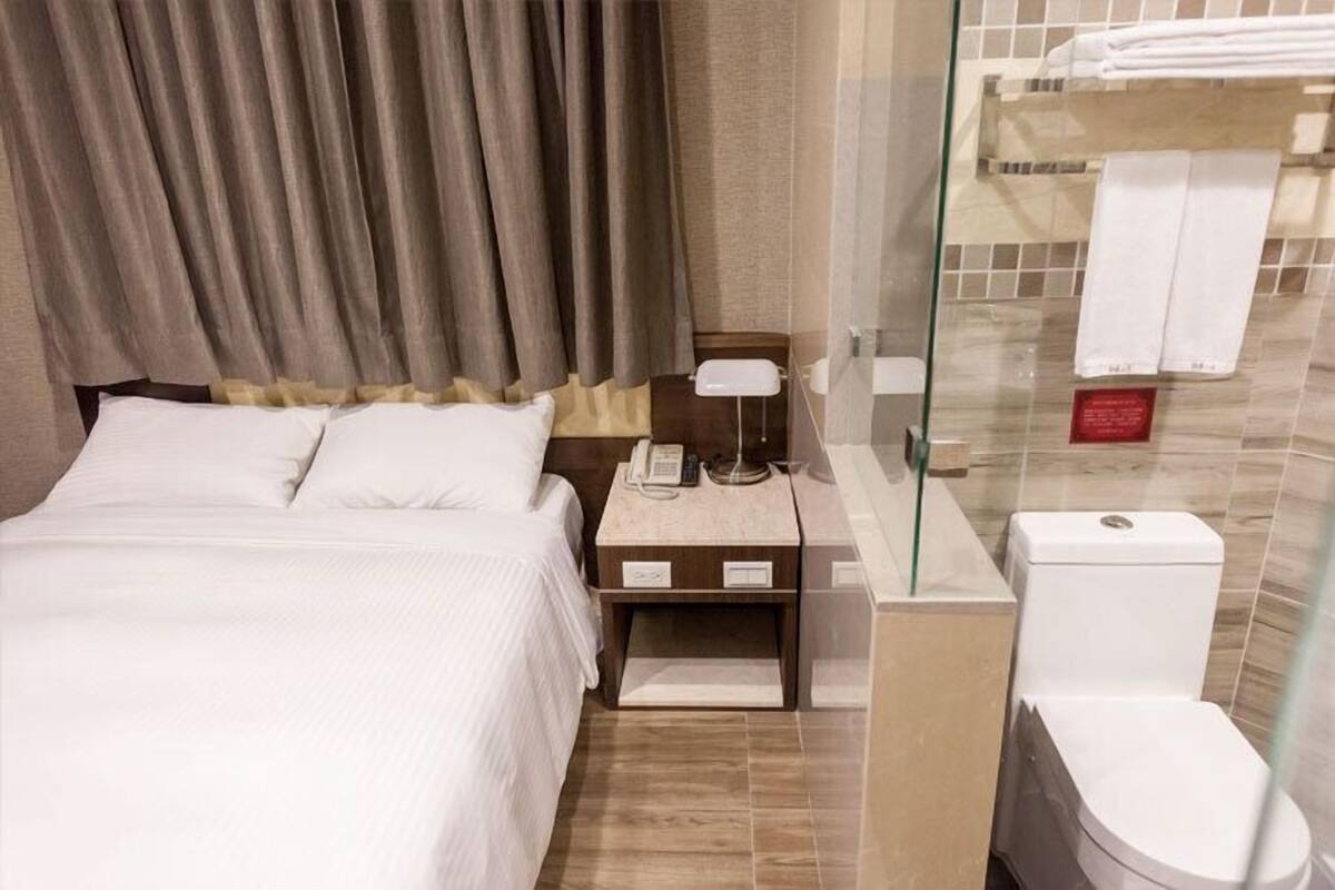 A hotel room with a bed, bedside table, lamp, and a glass-enclosed shower area with a toilet.