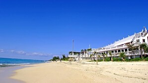 Un hotel frente a la playa con varios balcones, palmeras y un cielo azul despejado.