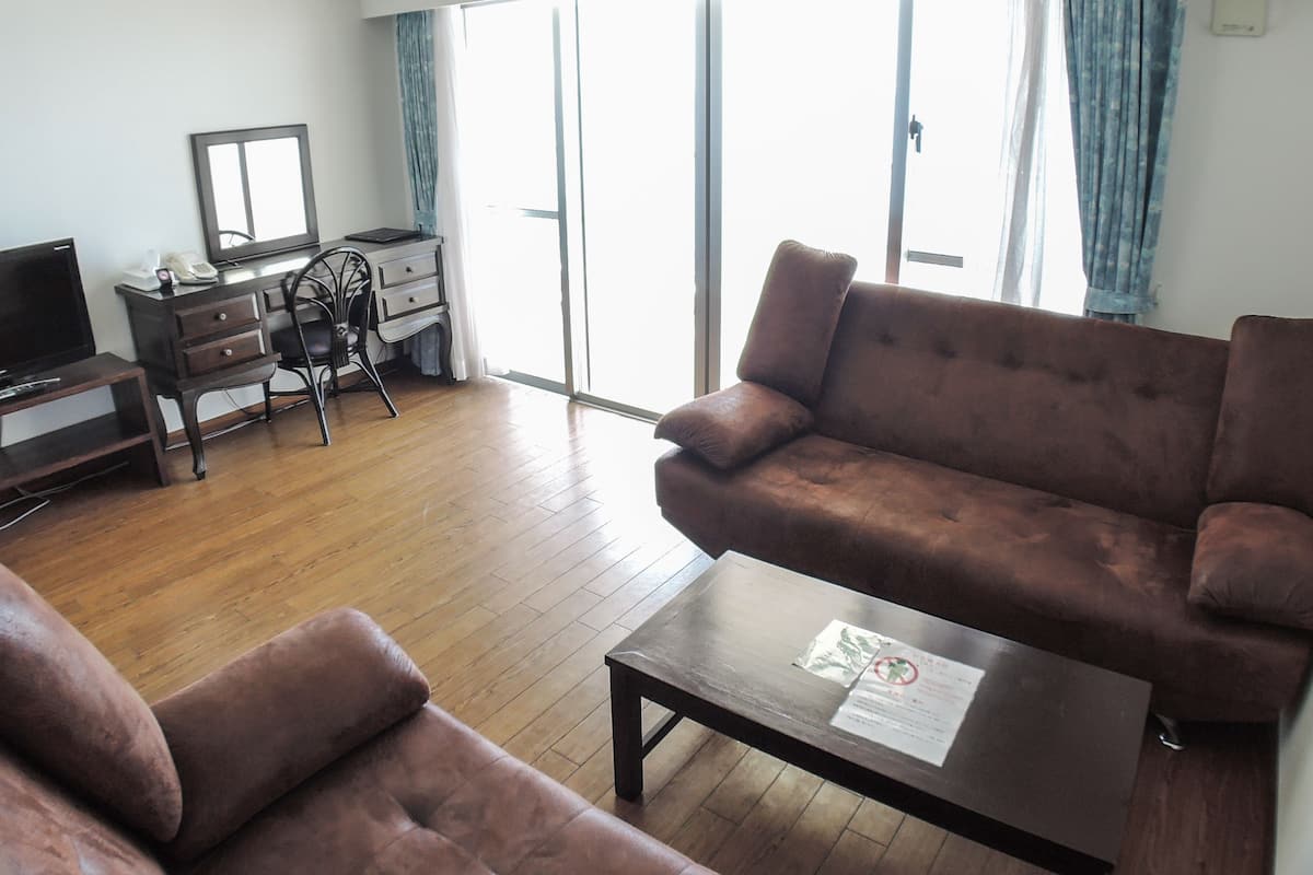 A living room with a sofa, a coffee table, a TV, a desk with a chair, and a sliding glass door leading to a balcony.