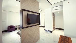 A modern hotel lobby with a large flat-screen TV mounted on a wooden panel, a purple armchair, a small round table, and a patterned rug on the floor. - De Ningrat Topas Galeria (Bandung)