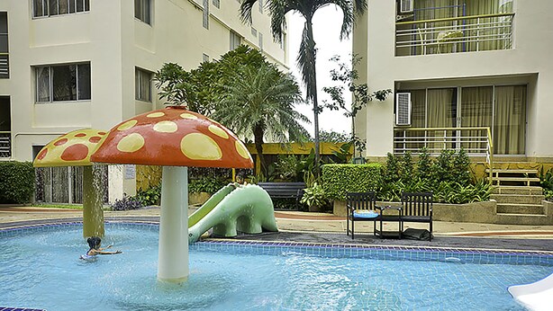 Una piscina con una gran fuente en forma de hongo, rodeada por un edificio con balcones y palmeras.