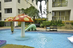 Una piscina con una gran fuente en forma de hongo, rodeada por un edificio con balcones y palmeras.
