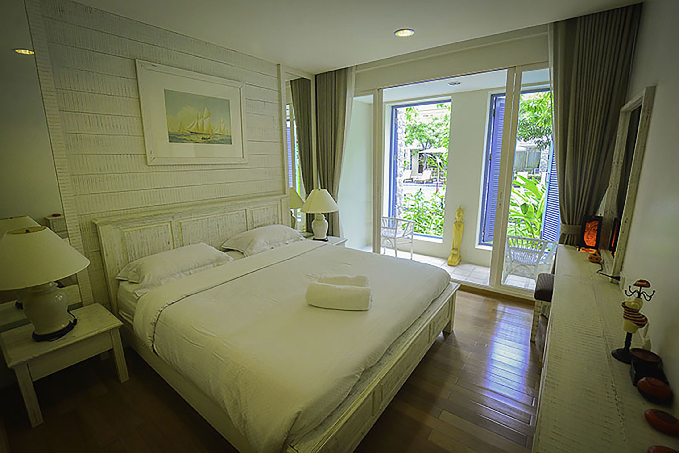 A spacious bedroom with a large bed, a nightstand, a lamp, and a window with a view of greenery.