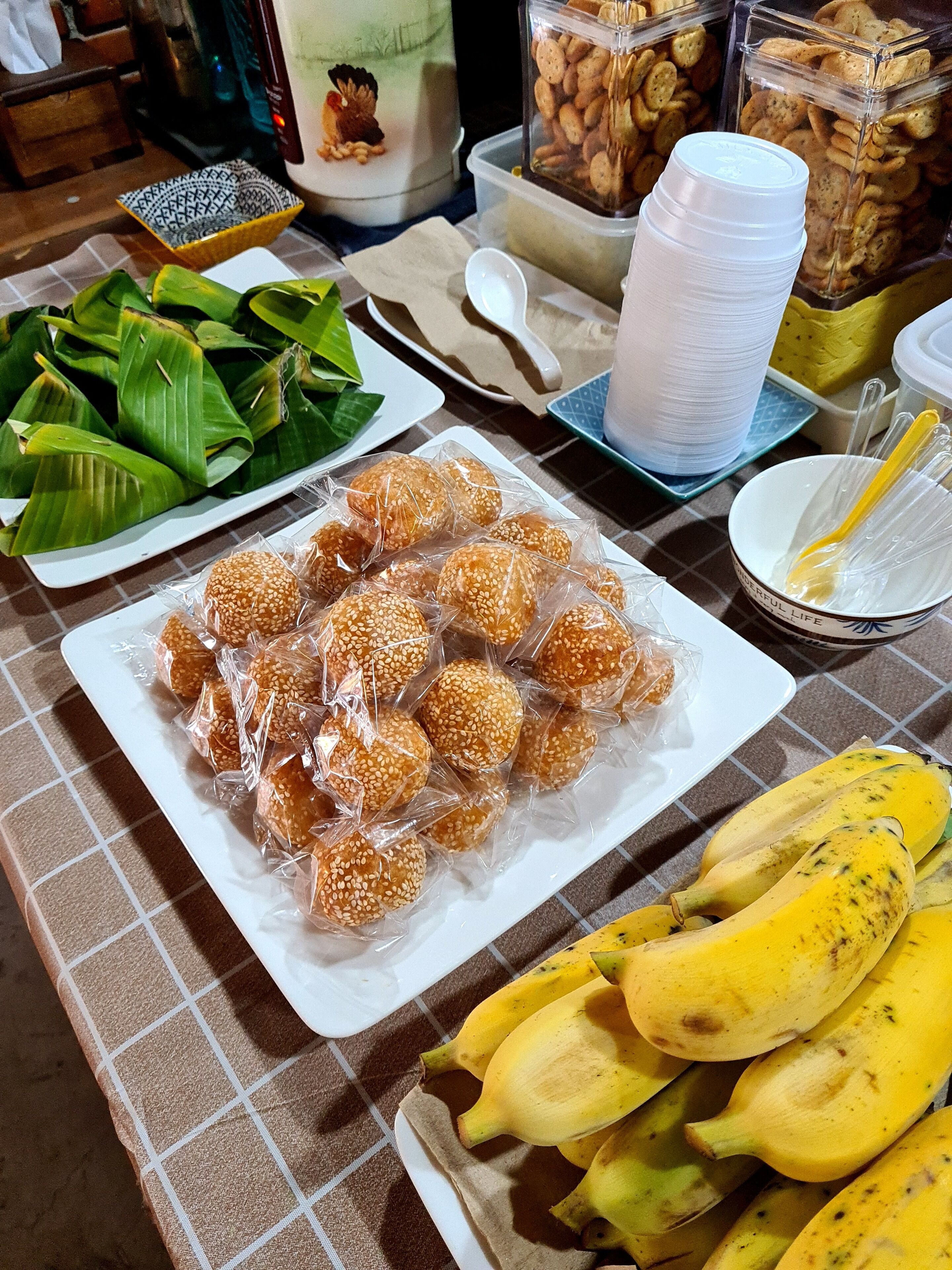 Una mesa con diversos alimentos, incluyendo un plato de bolitas de sésamo, un tazón de plátanos y recipientes con bocadillos.