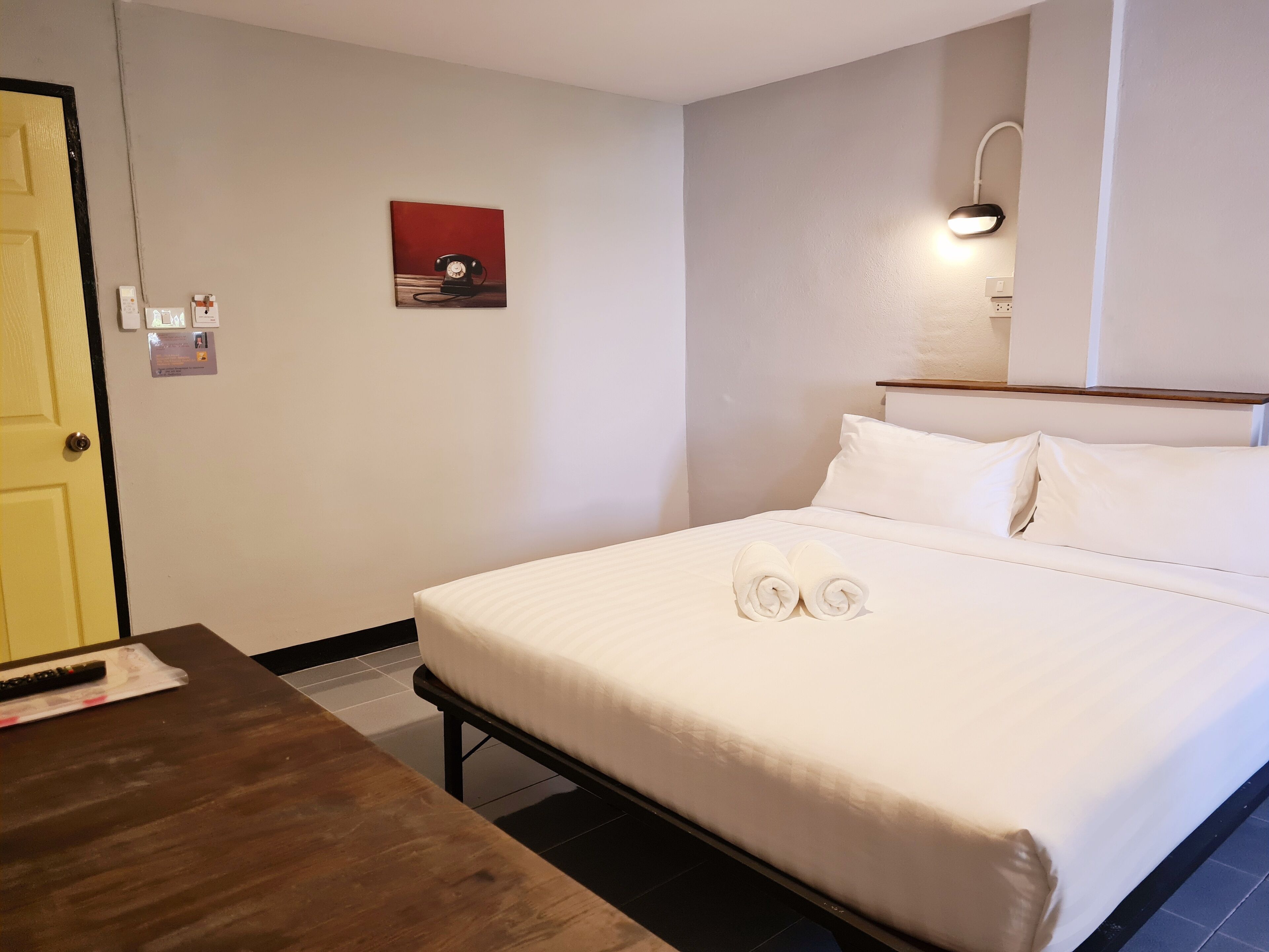 A hotel room with a neatly made bed, a bedside table, a wall-mounted lamp, and a painting on the wall.