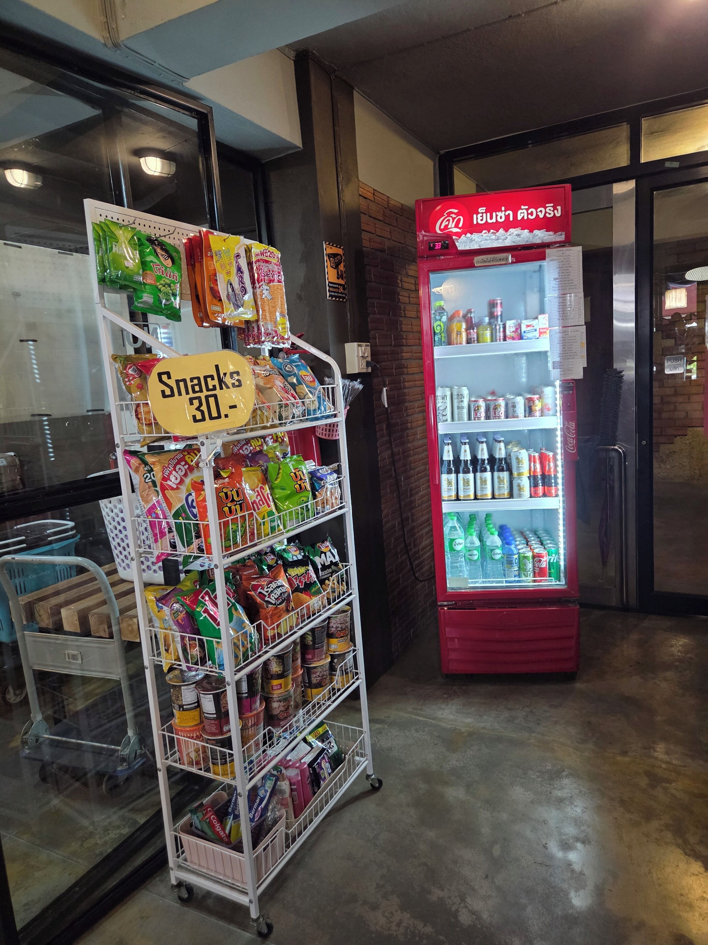 A snack display with a sign indicating a 30% discount, a Coca-Cola refrigerator stocked with beverages, and a metal cart with various items.