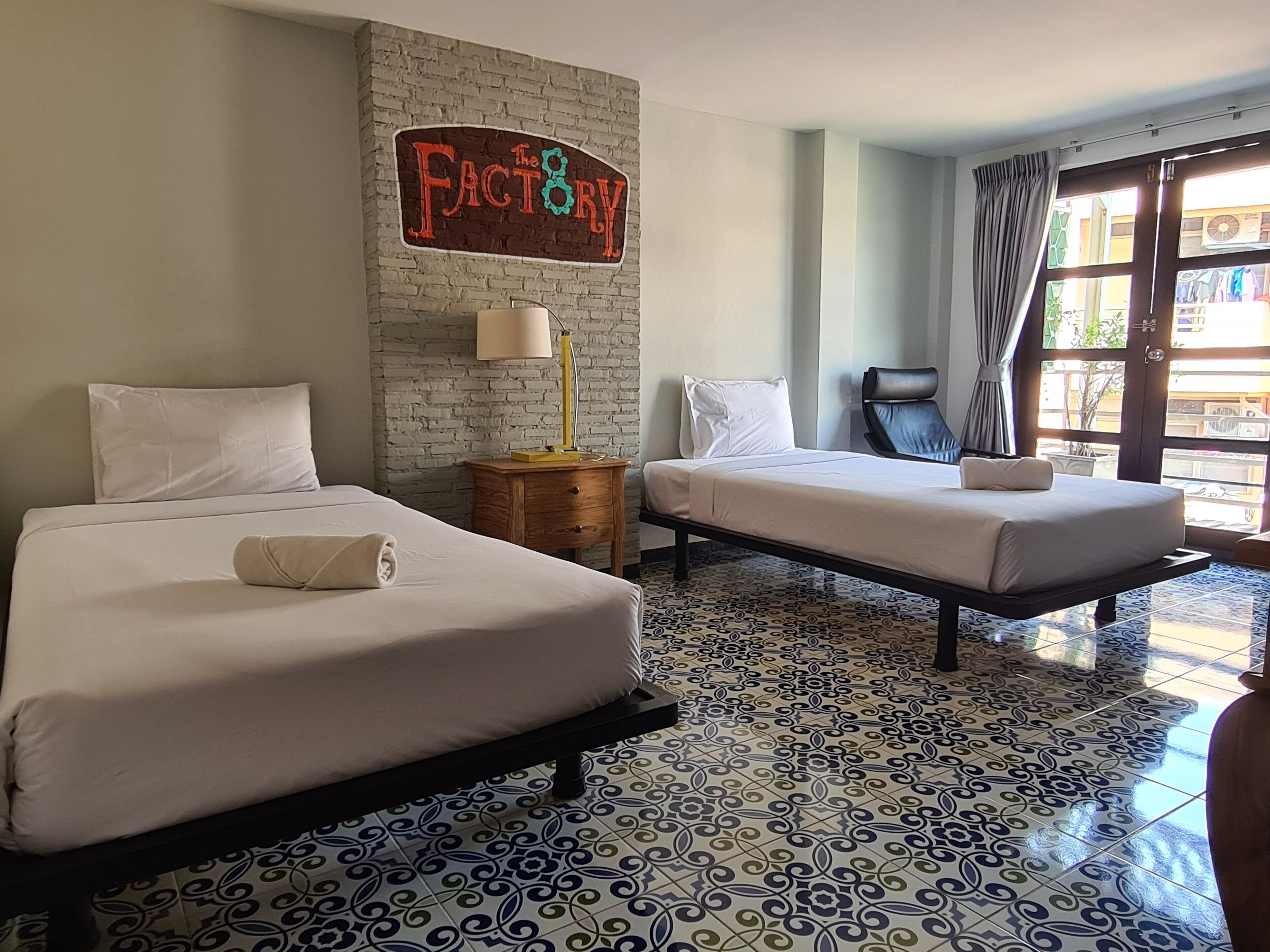 Habitación de hotel con dos camas, una pared de ladrillo con un letrero, una lámpara y vistas al exterior a través de una ventana.