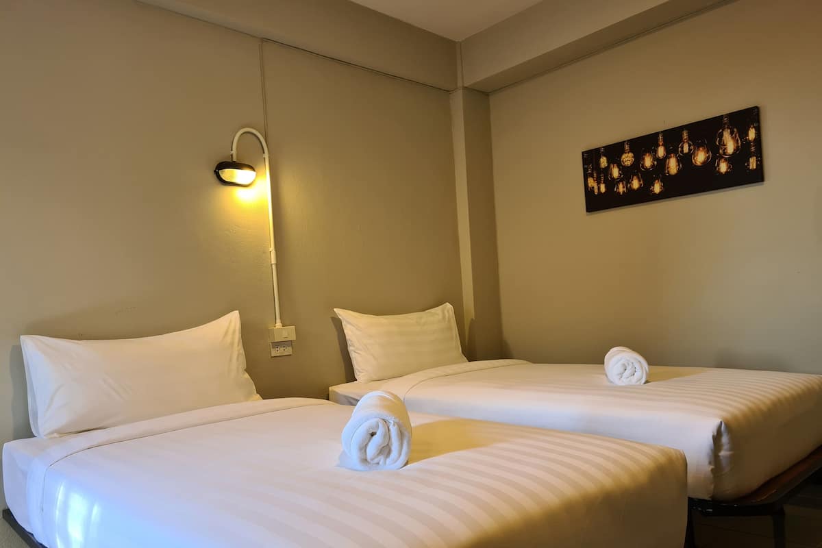 Une chambre d’hôtel avec deux lits, chacun équipé de draps blancs et d’une serviette enroulée. On y trouve un luminaire fixé au mur et un tableau représentant des ampoules.