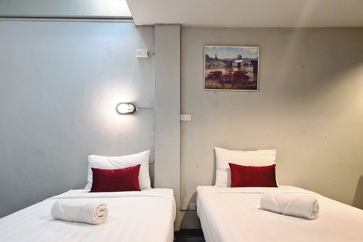 Une chambre d’hôtel avec deux lits, chacun doté de draps blancs et d’oreillers rouges, un tableau au mur et un interrupteur.