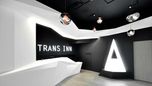 A modern hotel reception area with a white reception desk, a large illuminated 'A' sign, and pendant lighting. - Trans Inn (Taichung)