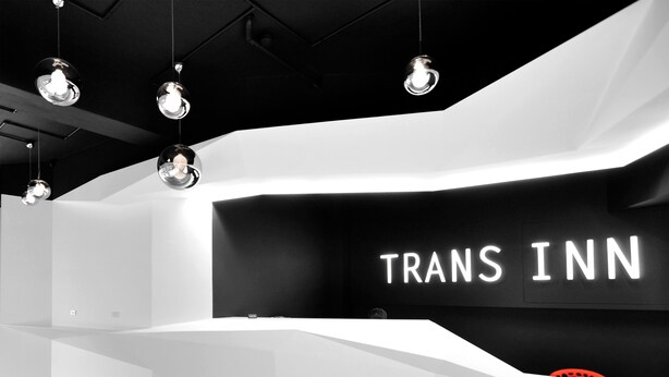 A modern hotel reception area with a curved white counter, a black wall with 'TRANS INN' signage, and hanging pendant lights. - Trans Inn (Taichung)