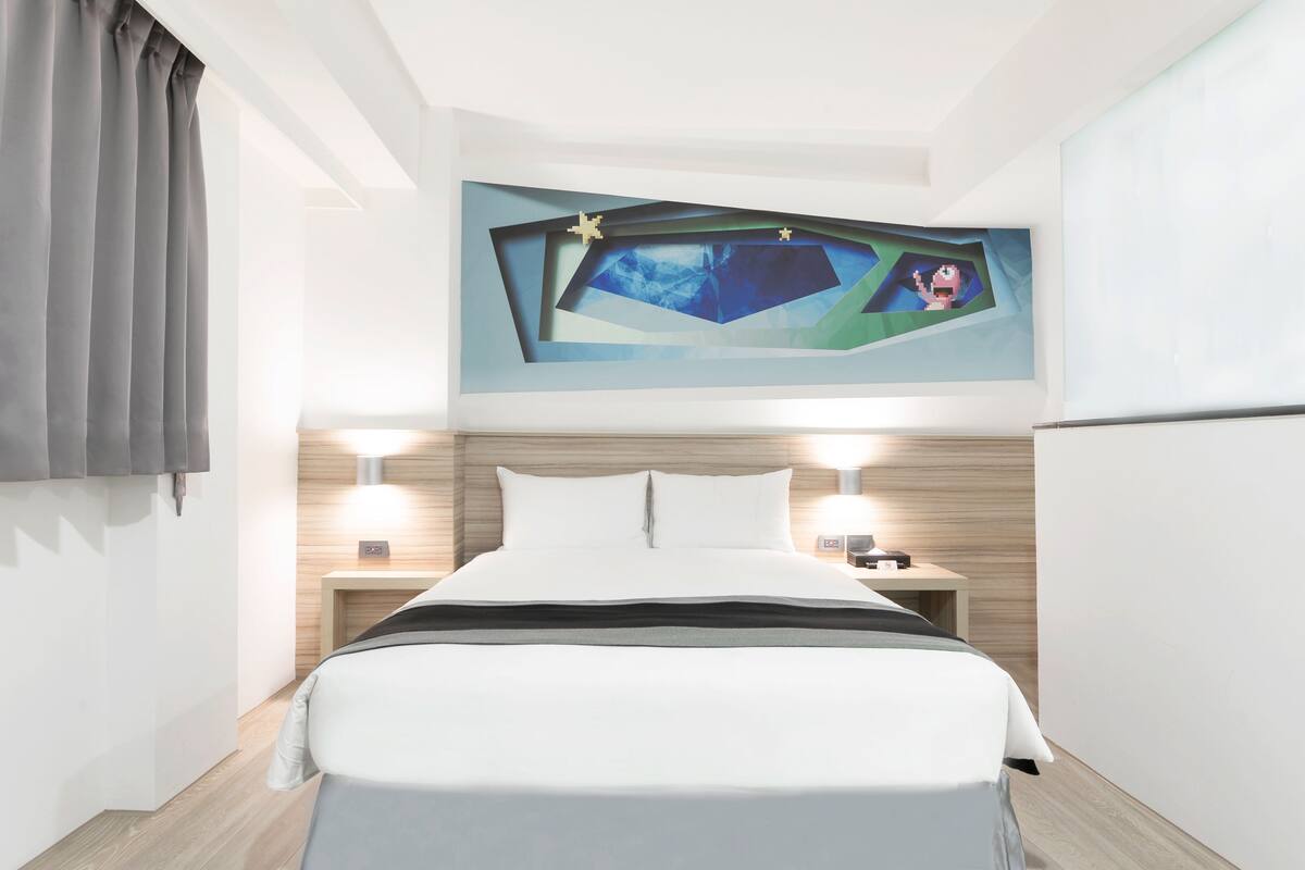 Una habitación de hotel moderna con una cama grande, dos mesitas de noche con lámparas, un cabecero de madera y una obra de arte colgada en la pared.