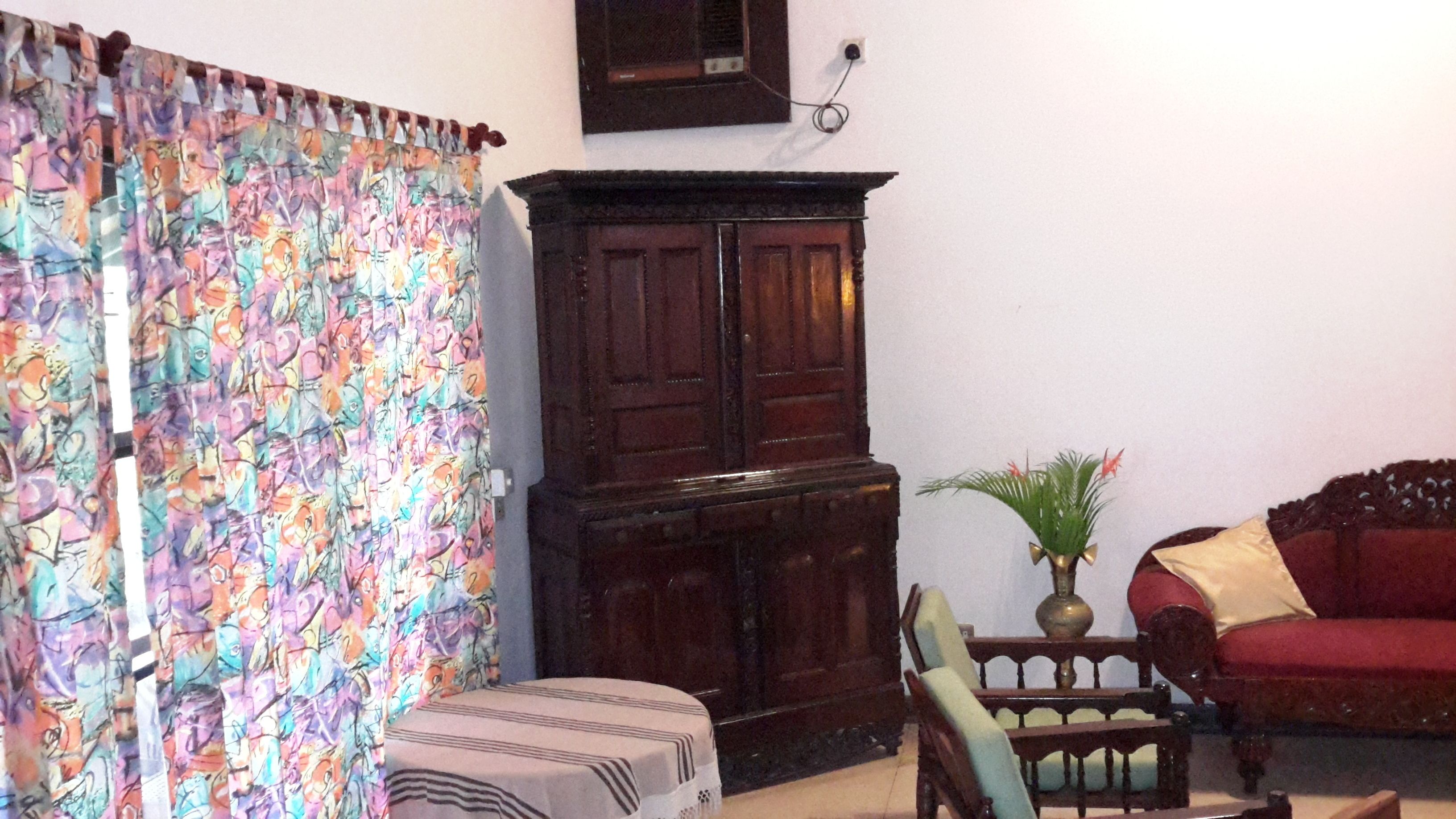 A room with a patterned curtain, a wooden cabinet, a red sofa, a green armchair, and a small table with a plant.