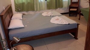A neatly made bed with pillows and a blanket, a bedside table with a lamp, and a chair in a room with a window and curtains.
