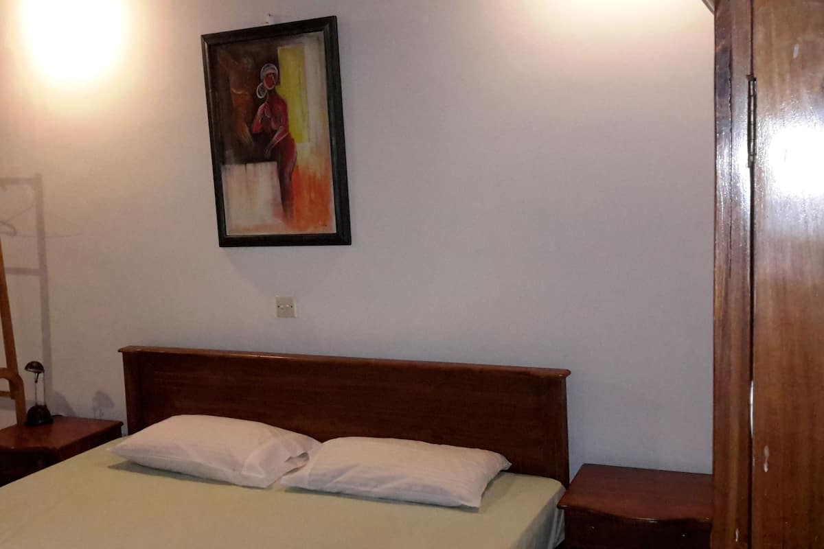 A bedroom with a wooden bed, white pillows, a wooden chair, a coat rack, and a painting on the wall.