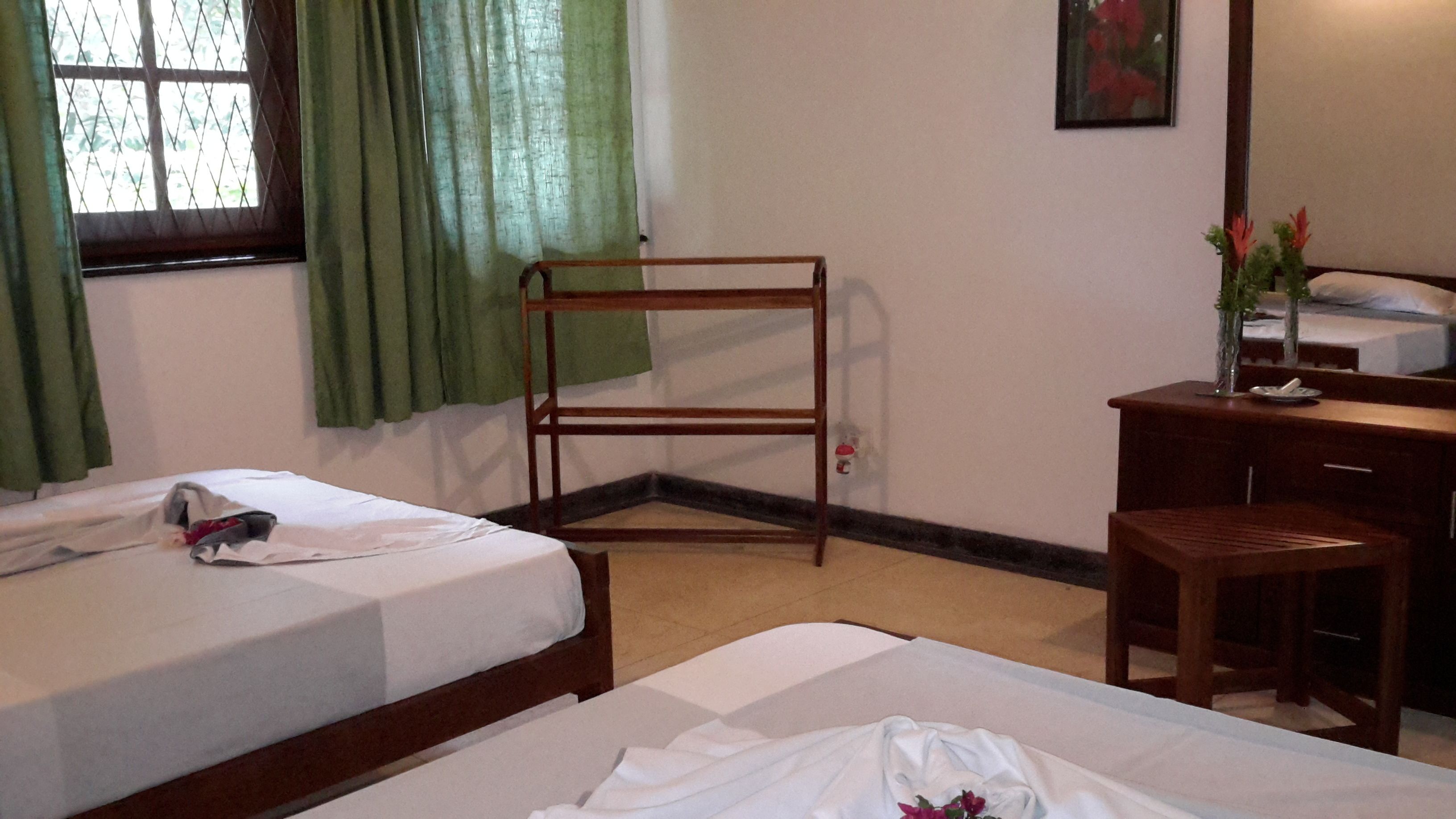 A hotel room with two beds, a wooden bench, a desk, a mirror, and a window with curtains.
