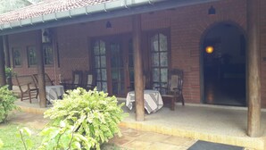 A covered outdoor seating area with brick walls, large windows, and a door leading to an interior space. - Arangala Bungalow (Naula)