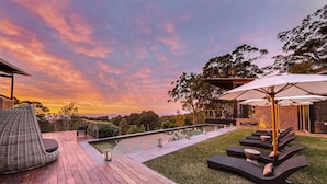 Eine Terrasse mit Liegestühlen und Sonnenschirmen, ein Schwimmbad und Blick auf den Sonnenuntergang.