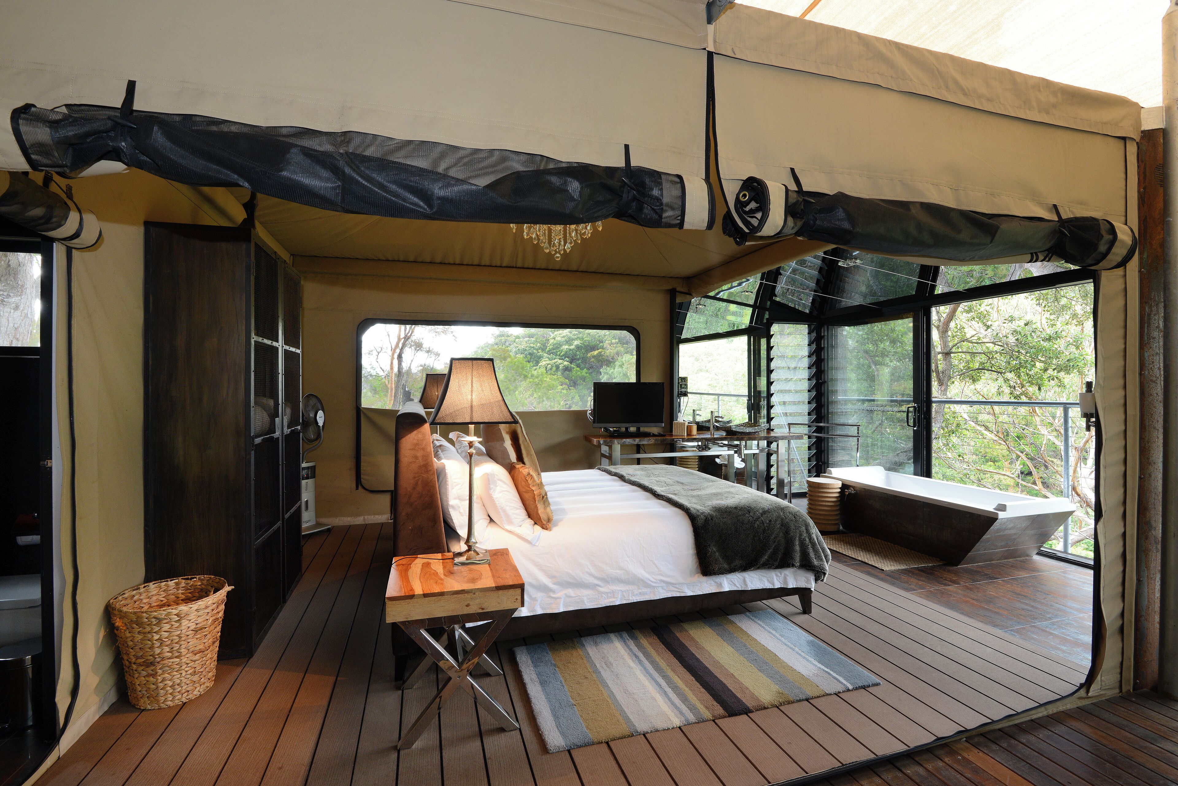 A tented room with a bed, a desk, a lamp, a television, and a view of the outdoors.