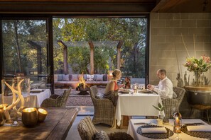 Un espace extérieur aménagé pour les repas, avec des meubles en osier, un foyer et une vue sur les arbres à travers de grandes fenêtres.