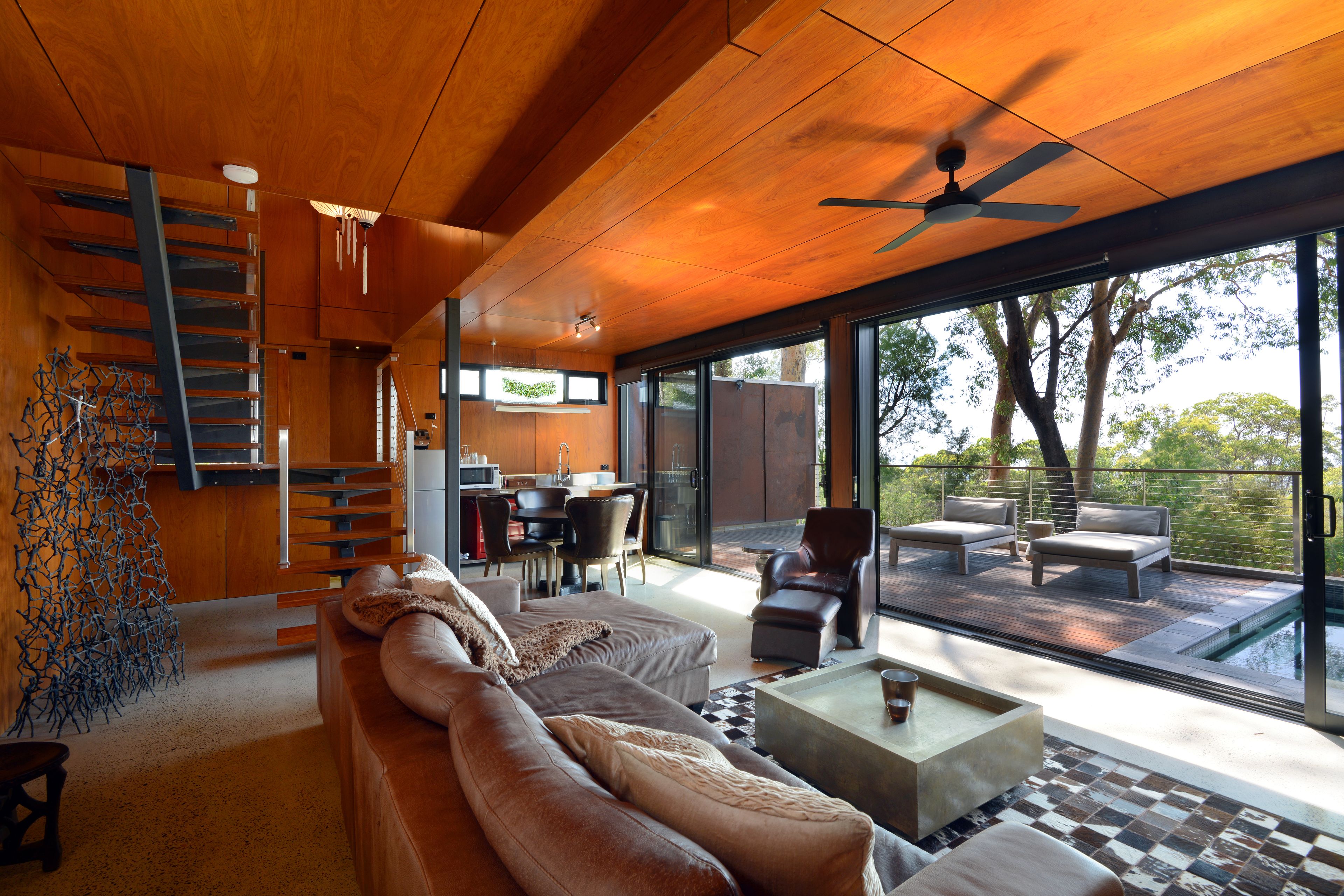 A modern living room with a wooden ceiling, a staircase, a dining area, and a view of a swimming pool.