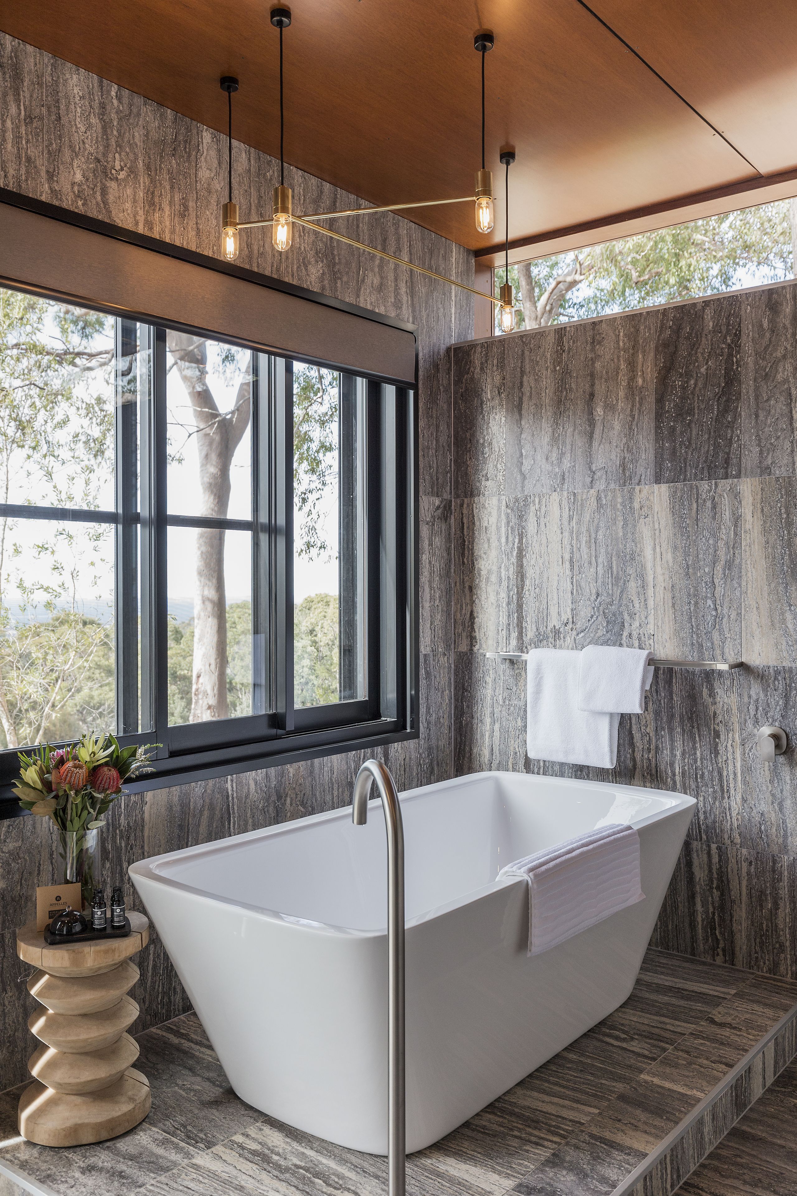 A modern bathroom with a freestanding bathtub, a large window with a view of trees, and a wooden side table with a vase.