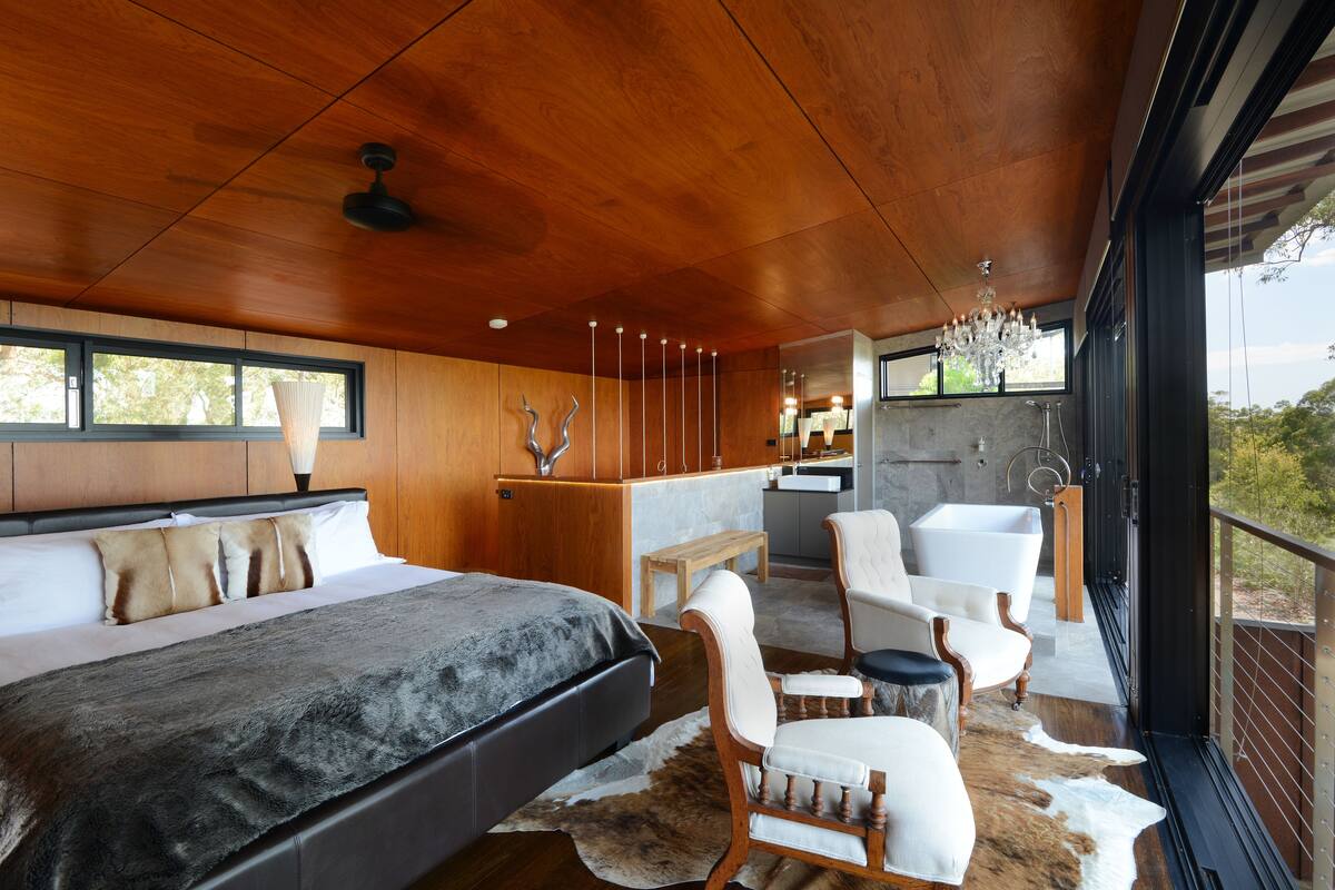 Une chambre à coucher moderne avec un grand lit, un coin salon comprenant un fauteuil et une petite table, un plafond en bois et une vue sur l’extérieur grâce à de grandes fenêtres.