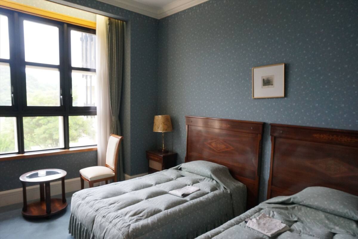 Ein Hotelzimmer mit zwei Betten, einem Sessel, einem kleinen Tisch und einem Fenster mit Vorhängen.