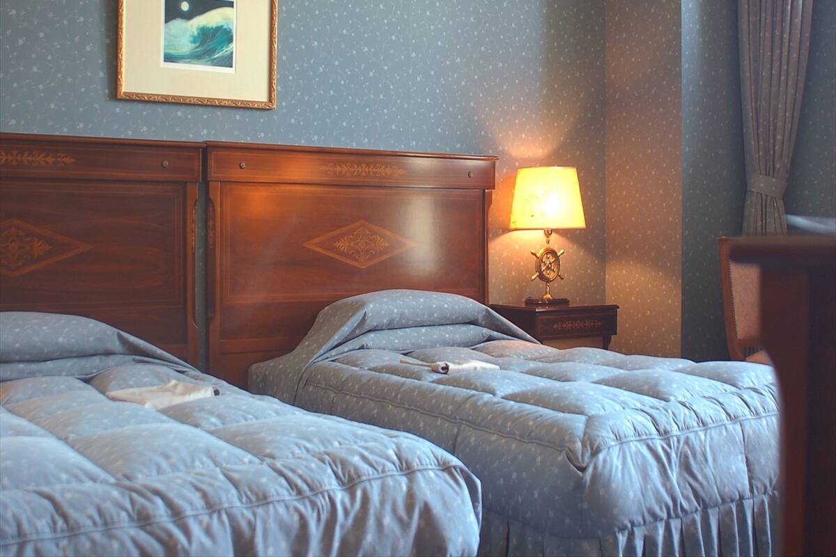 Ein Zimmer mit zwei Betten, einem Nachttisch mit Lampe, einem gerahmten Bild an der Wand und einem Wandpapier mit Muster.