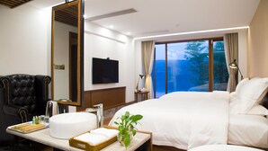 A hotel room with a large bed, a sitting area with a chair, a vanity with a mirror, a television, and a window with a view of trees. - Fishman homestay (Hangzhou)