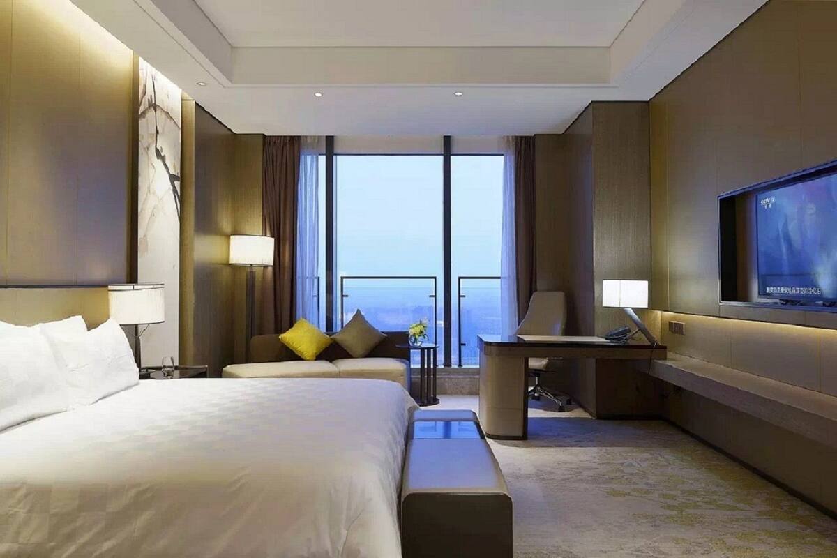 A hotel room with a large bed, a desk, a chair, a TV, and a view of the city through large windows.