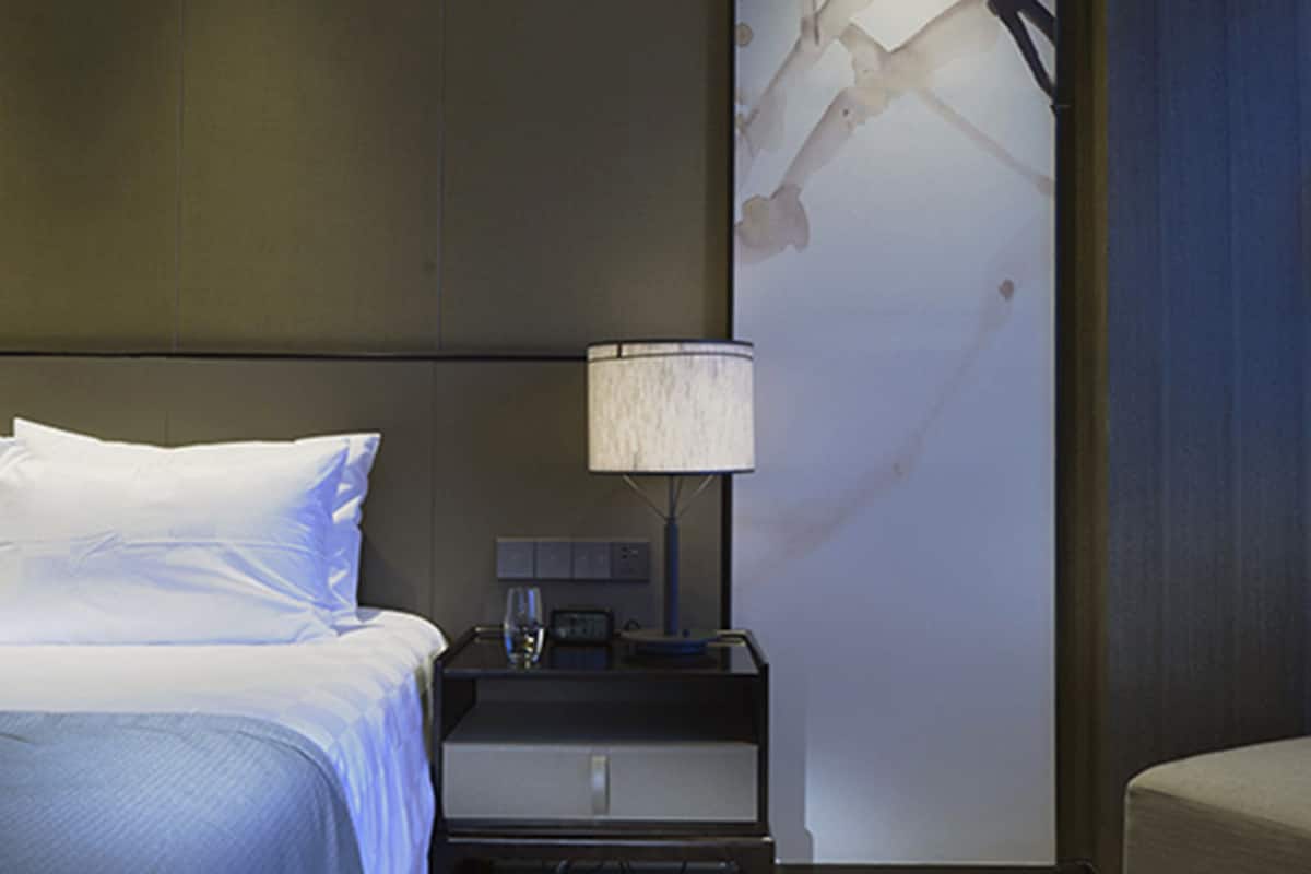 A hotel room with a bed, bedside table, lamp, and a wall with a branch artwork.