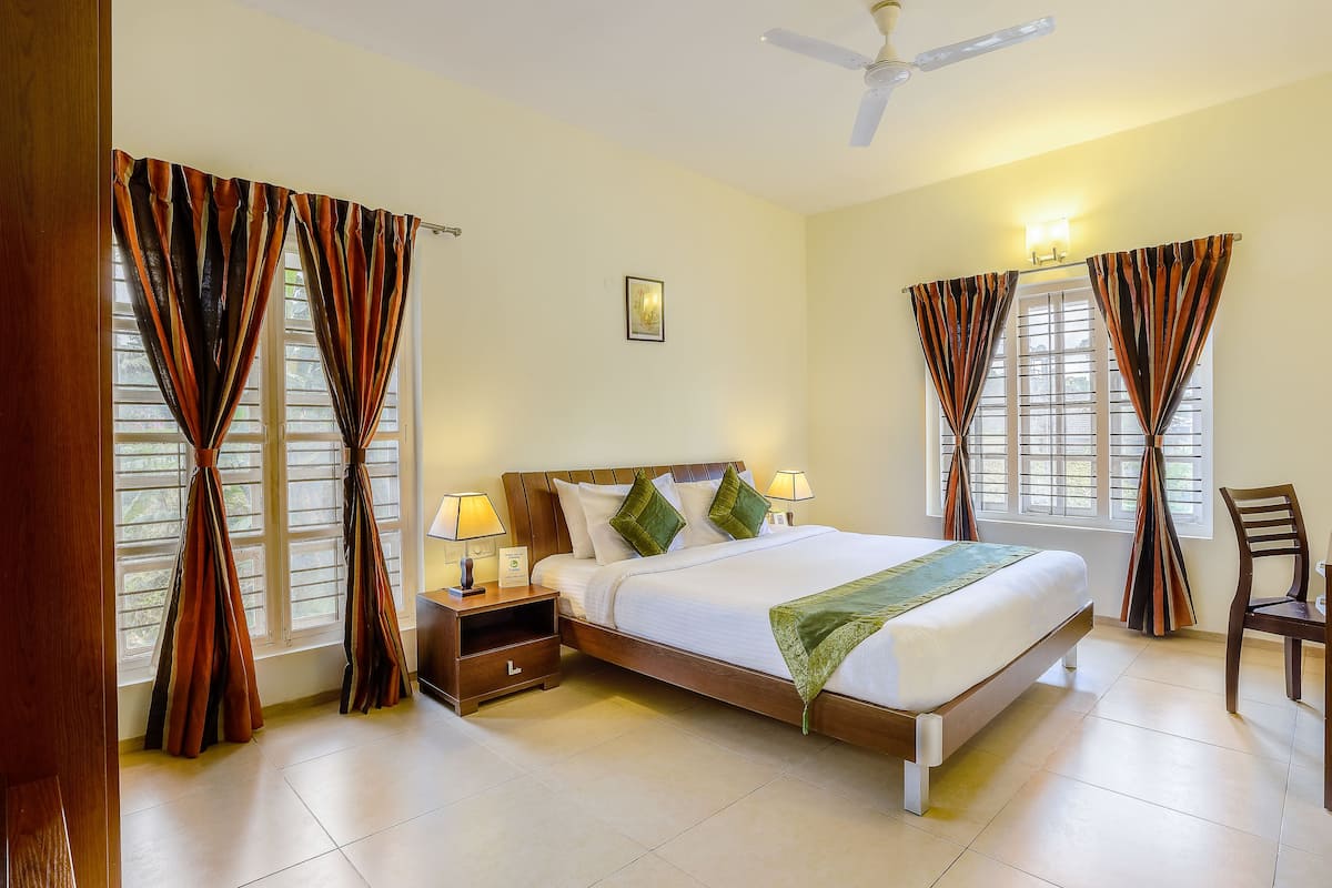 Quarto de hotel bem arrumado, com cama, duas mesas de cabeceira com abajures, uma cadeira e um ventilador de teto.
