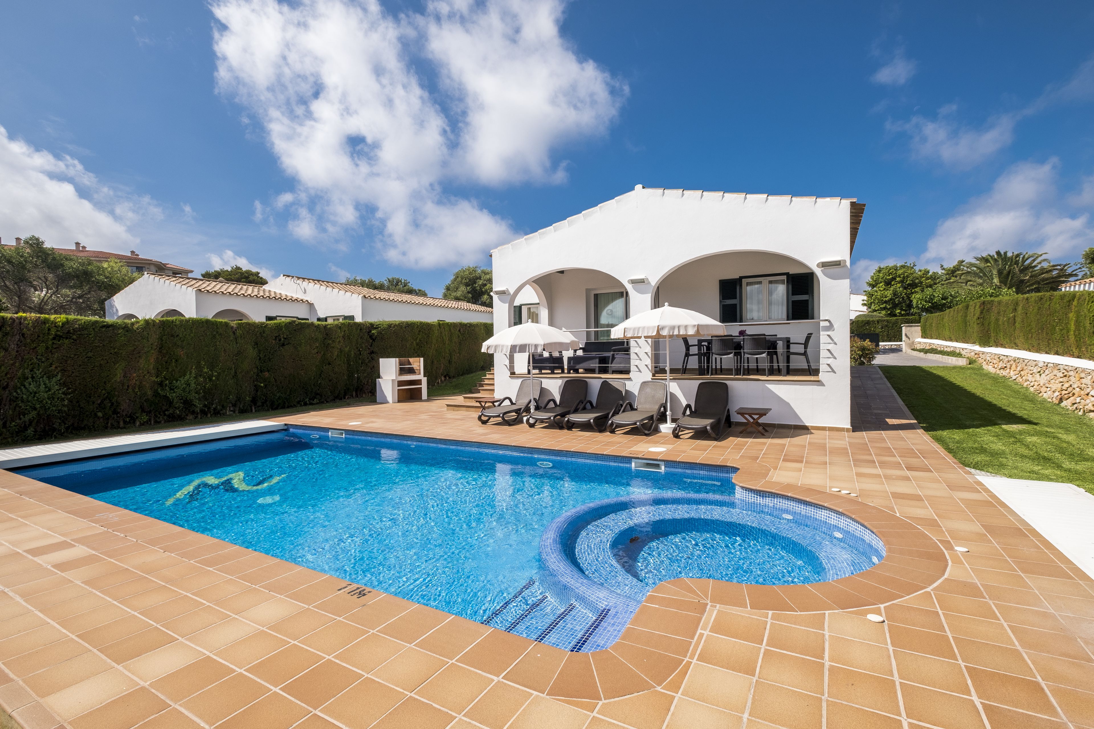 A swimming pool with a tiled deck, a covered outdoor seating area, and a white building with arches.