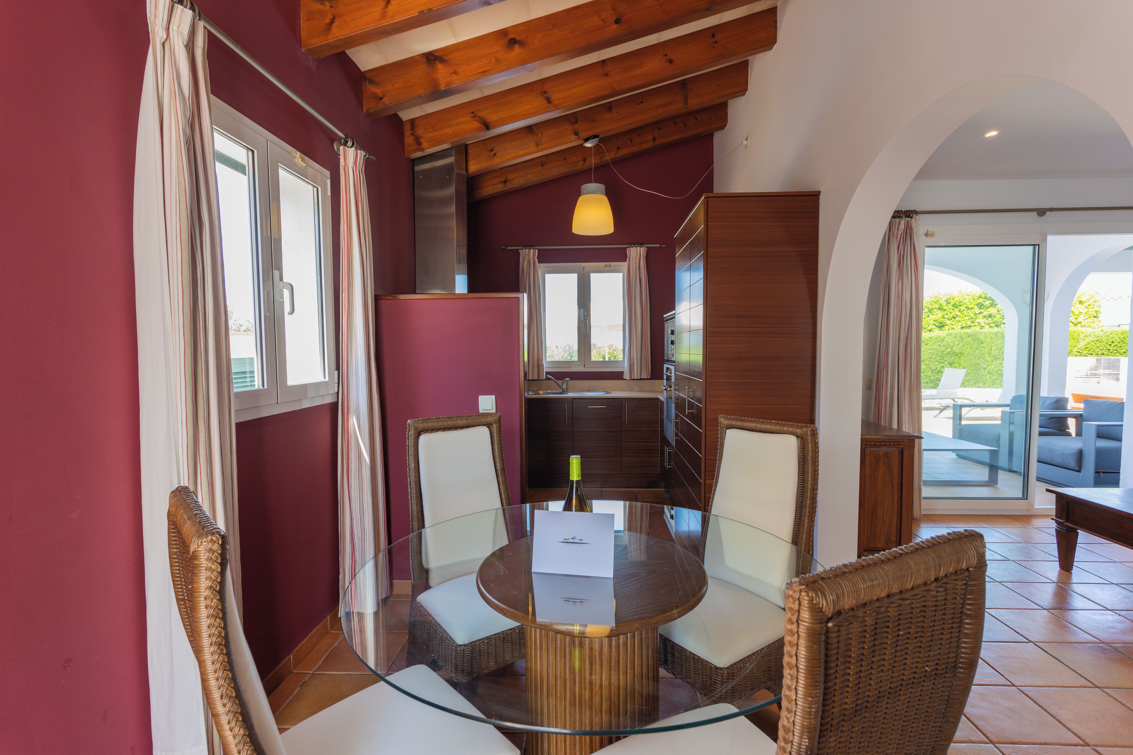 Un comedor con una mesa de vidrio rodeada de sillas de mimbre, techo de madera y vista al patio exterior.