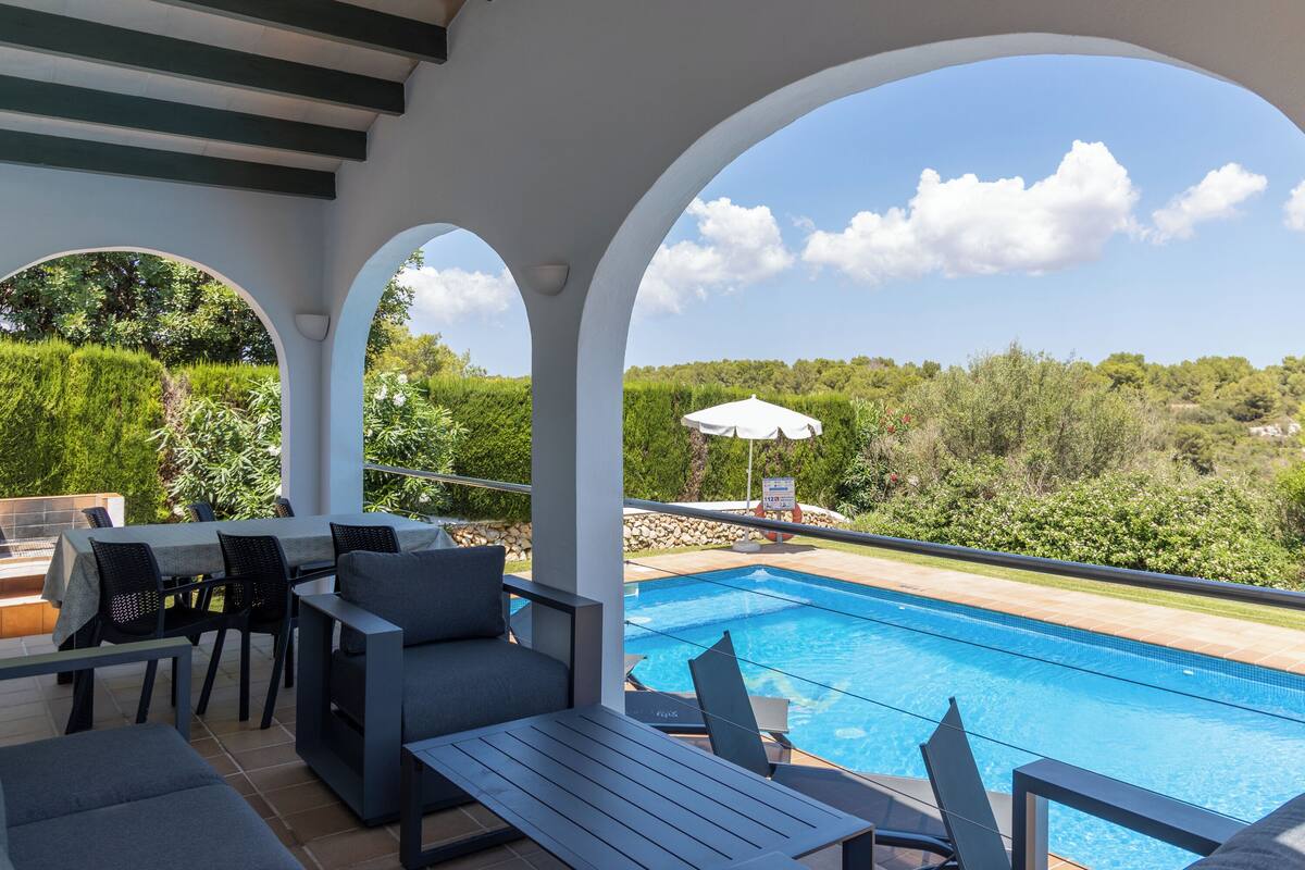 Zona de piscina al aire libre con comedor y vistas al entorno verde.