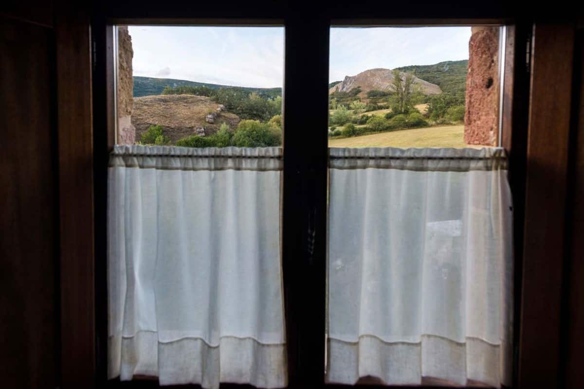 A window with white curtains, offering a view of a mountainous landscape with greenery and rocky outcrops.