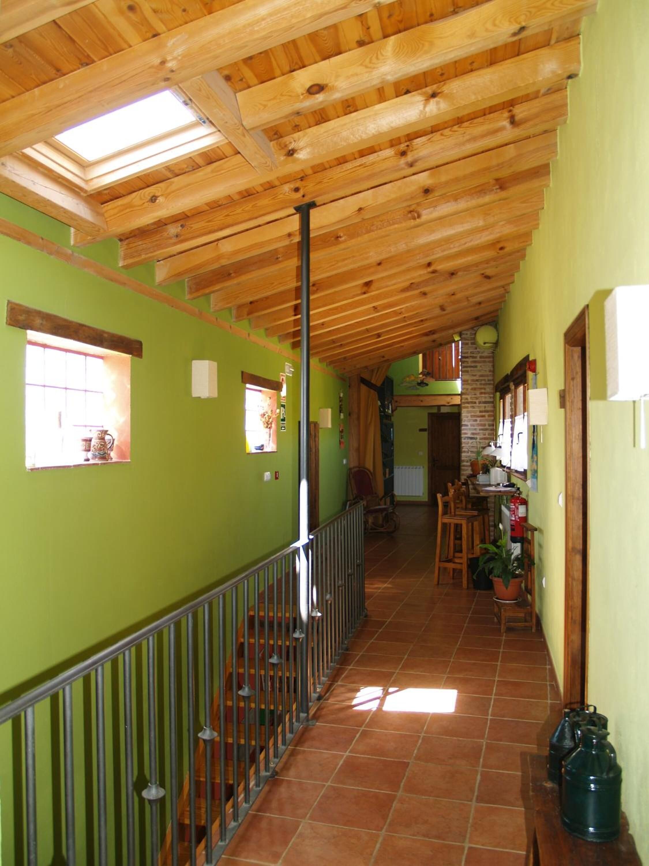 A corridor with a wooden ceiling, green walls, and a metal railing. There are windows, a staircase, and a brick wall with a door.