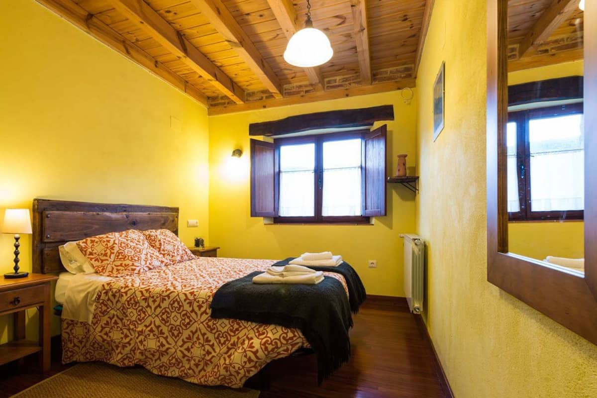 A room with a wooden ceiling, a bed with patterned bedding, a nightstand with a lamp, and a window with a view.