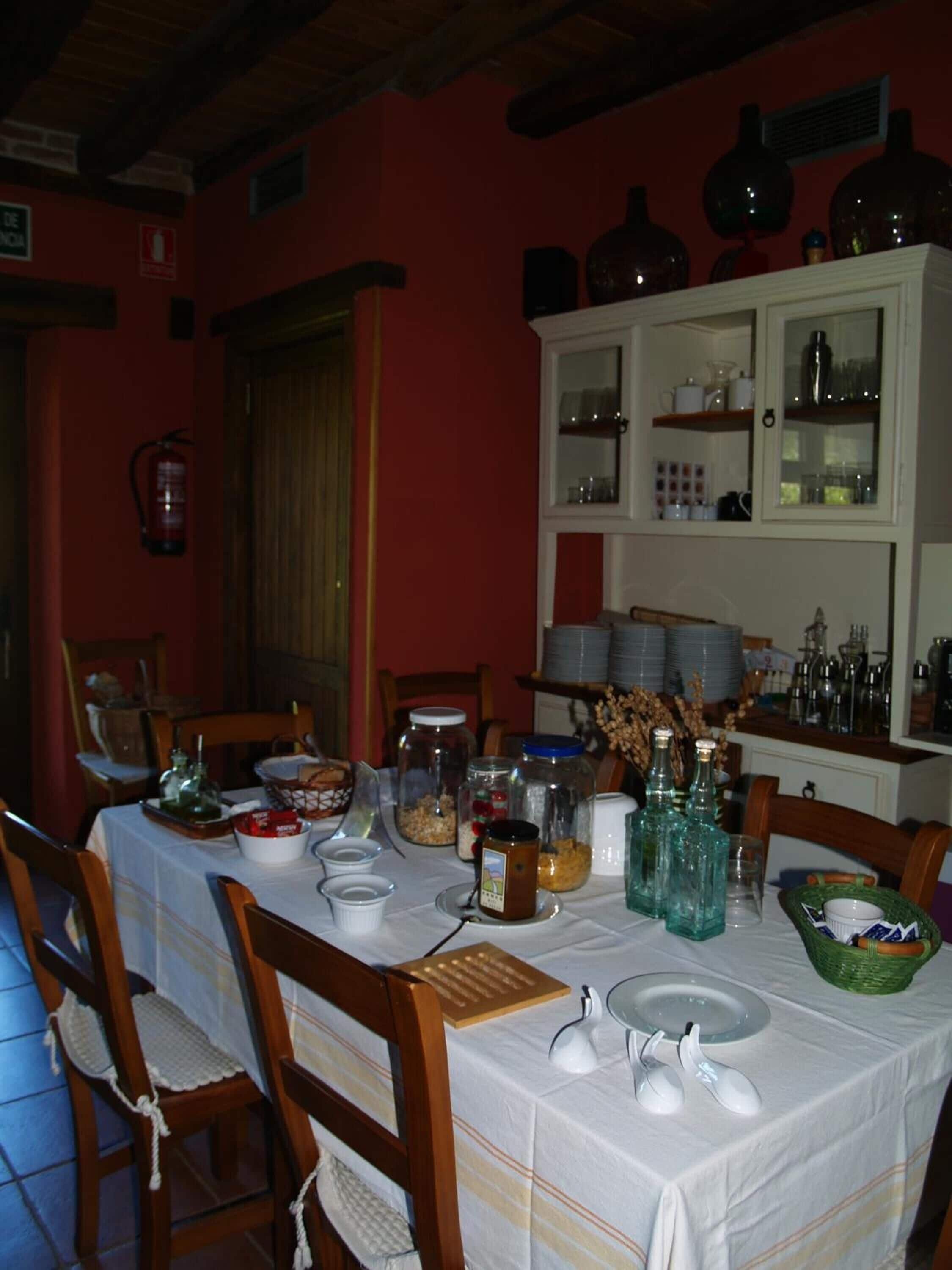 Un comedor con una mesa dispuesta para cuatro personas, rodeada de sillas de madera. Se observa un armario blanco con puertas de vidrio al fondo, que contiene varios utensilios de cocina.