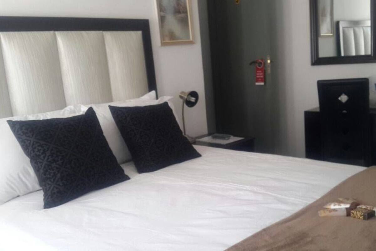 A hotel room with a large bed, two black patterned pillows, a wooden nightstand, a mirror, and a small table with a book and a remote control.