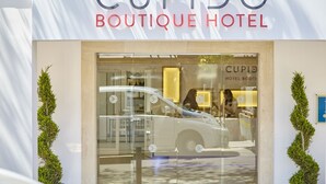 El exterior del CUPIDO Boutique Hotel, con un diseño moderno, entrada de vidrio y dos plantas verdes simétricas a cada lado.