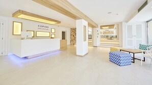 Un área de recepción de hotel moderna con un mostrador blanco, una pared de acento de piedra y una zona de estar con un pouf estampado y una mesa de madera.