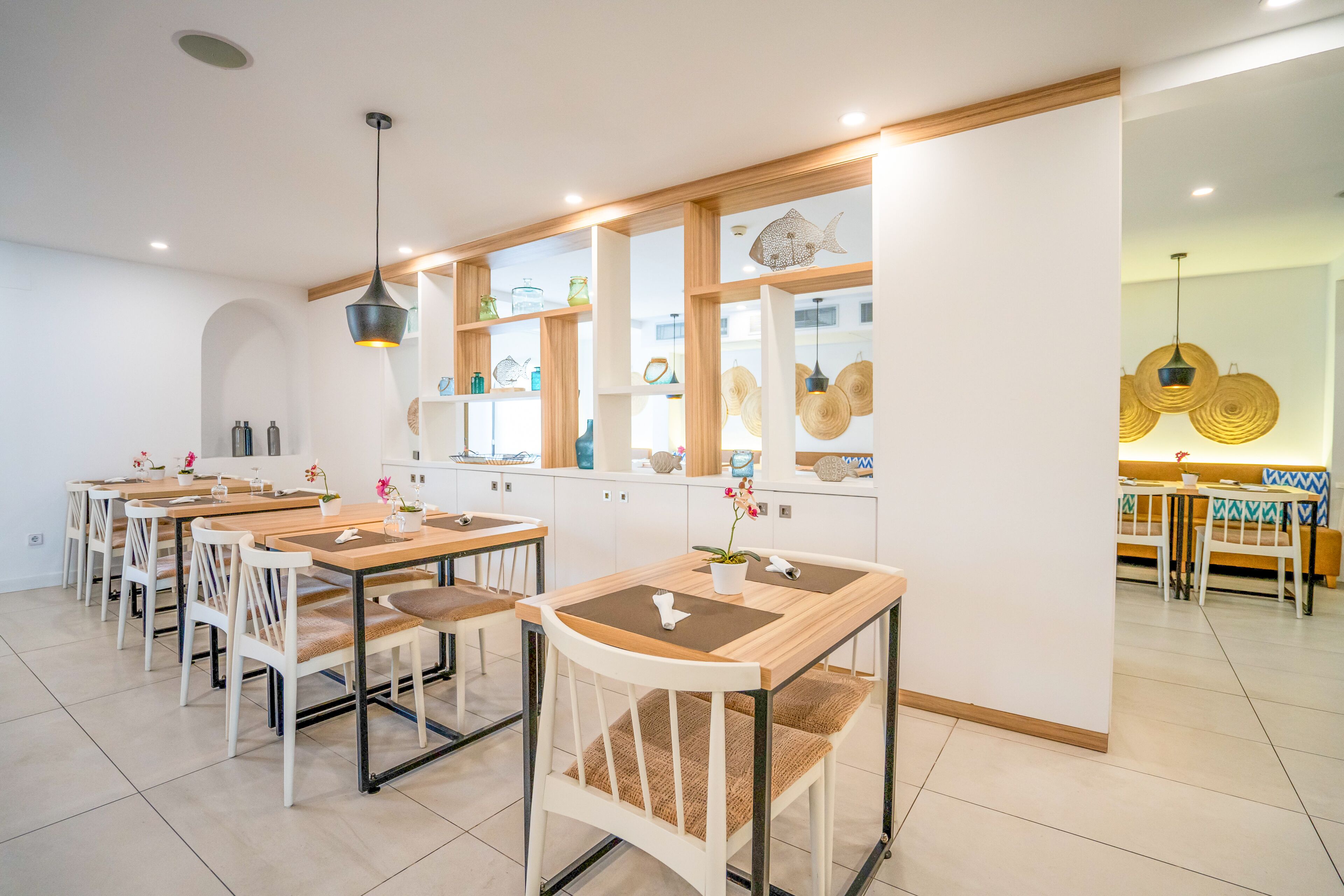 Un comedor moderno con varias mesas y sillas, una zona de cocina con estantes que exhiben diversos objetos, y un área de estar con un sofá y una mesa.