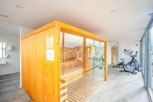 A wooden sauna with glass walls, a wooden bench, and exercise bikes in a modern room with tiled flooring.