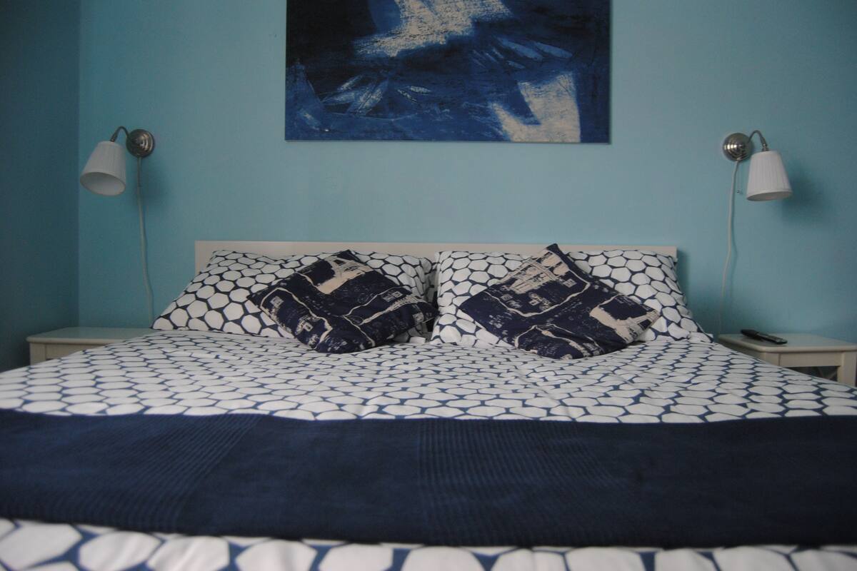 A bed with a patterned bedspread and two matching pillows, a wall-mounted abstract painting, and two bedside lamps.