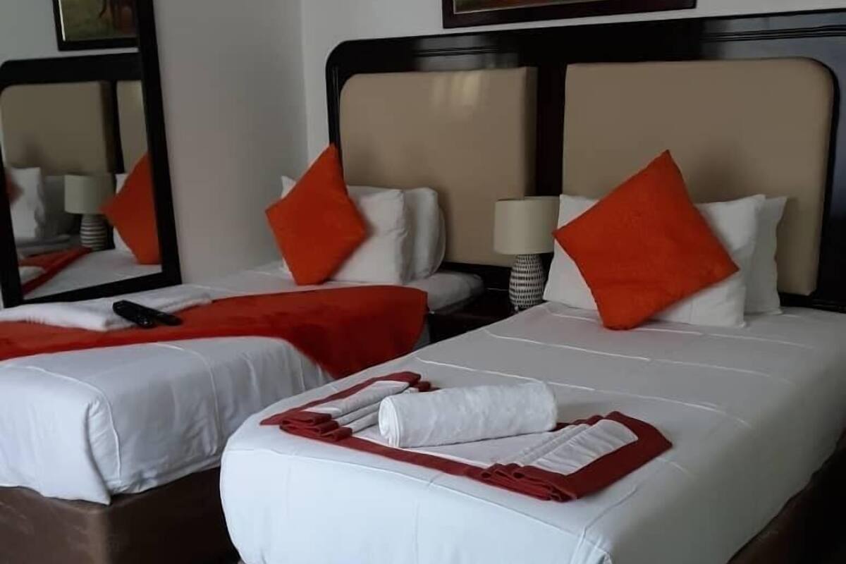 A hotel room with two beds, each with white bedding and red pillows. There are two bedside tables with lamps, and a large mirror on the wall.